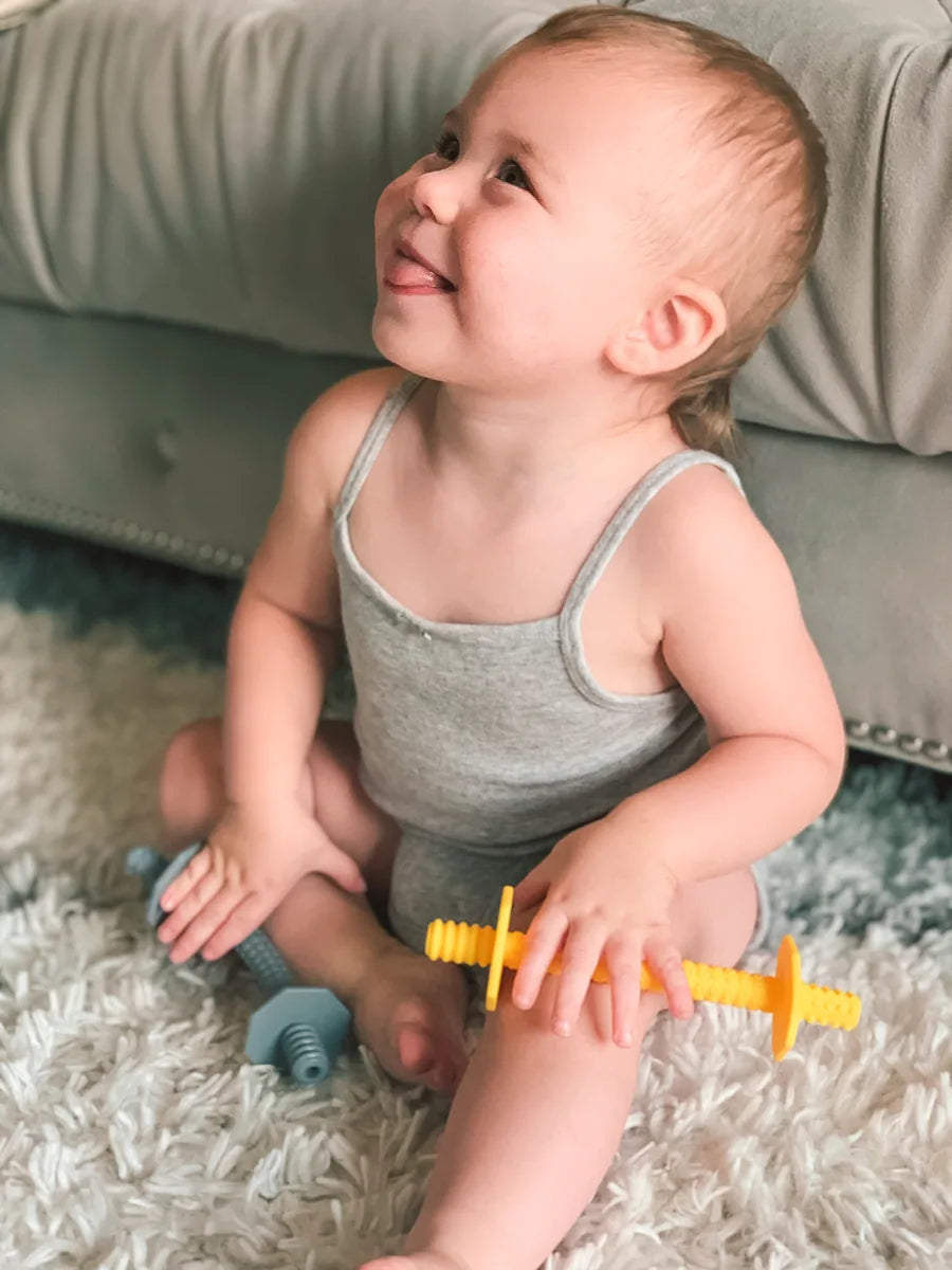 Smiling baby sitting on a rug holding two Lull Kids teething straws